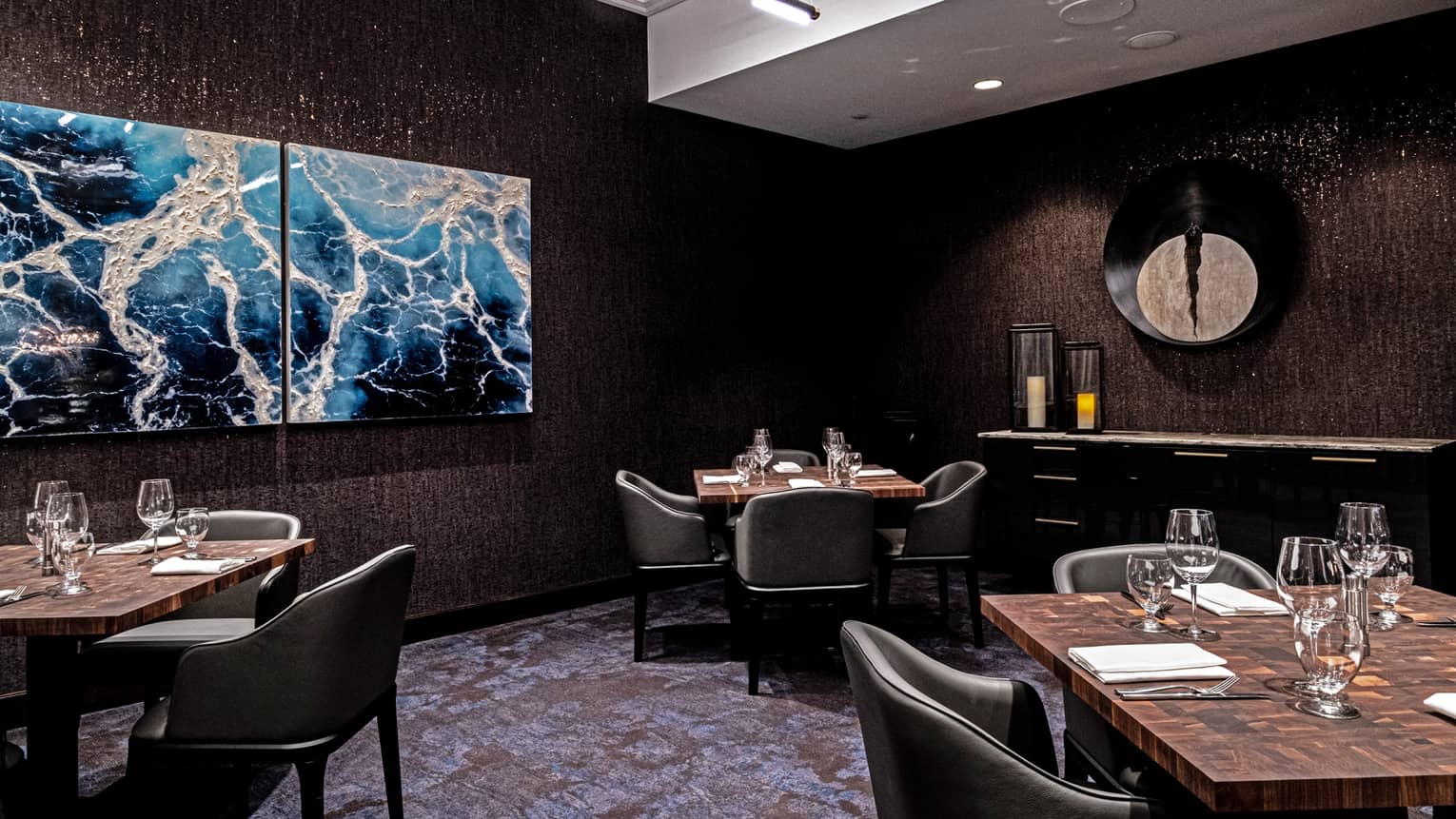An upscale restaurant with dark walls and abstract art, three tables each set for four with leather club chairs.