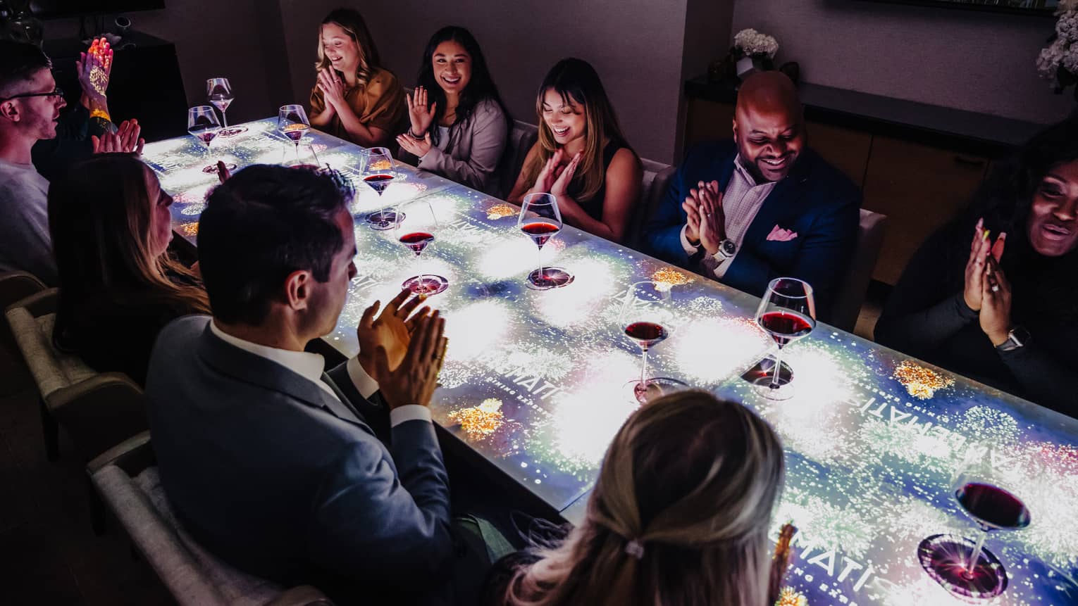A large group of people sitting at a table with a screen on top of it drinking wine.