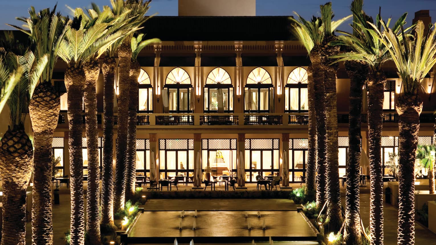 Fountains between rows of tall palm trees in front of illuminated Four Seasons Resort Marrakech balconies at night