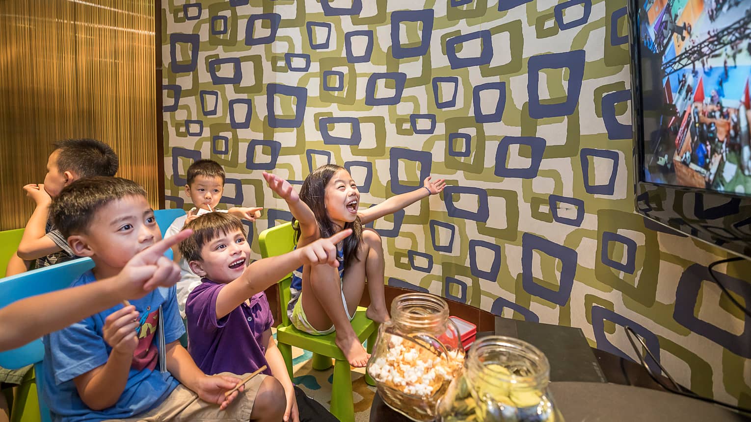 Children sit on chairs in Kids Club, laugh and point at TV screen