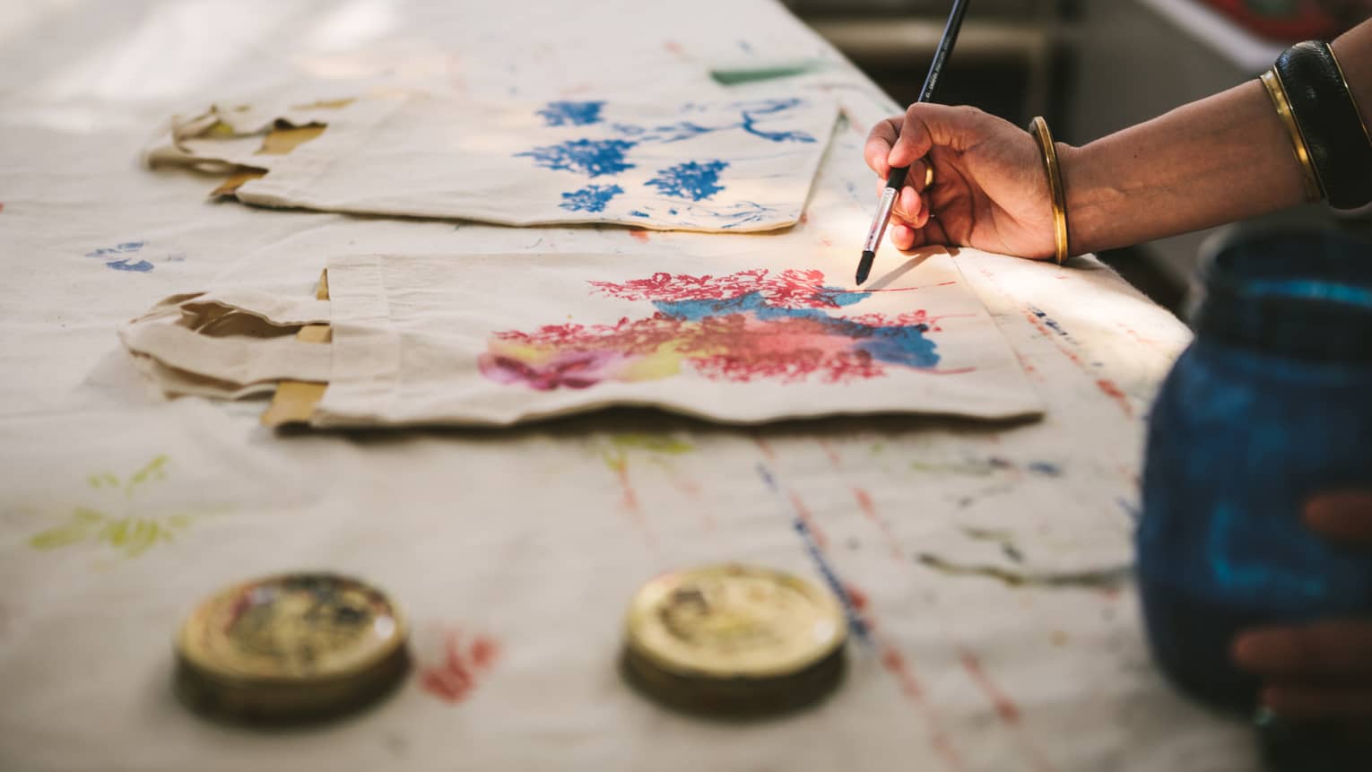 Close-up of a gold-bangled hand wielding a long brush to paint an intricate pattern on a cloth tote bag.