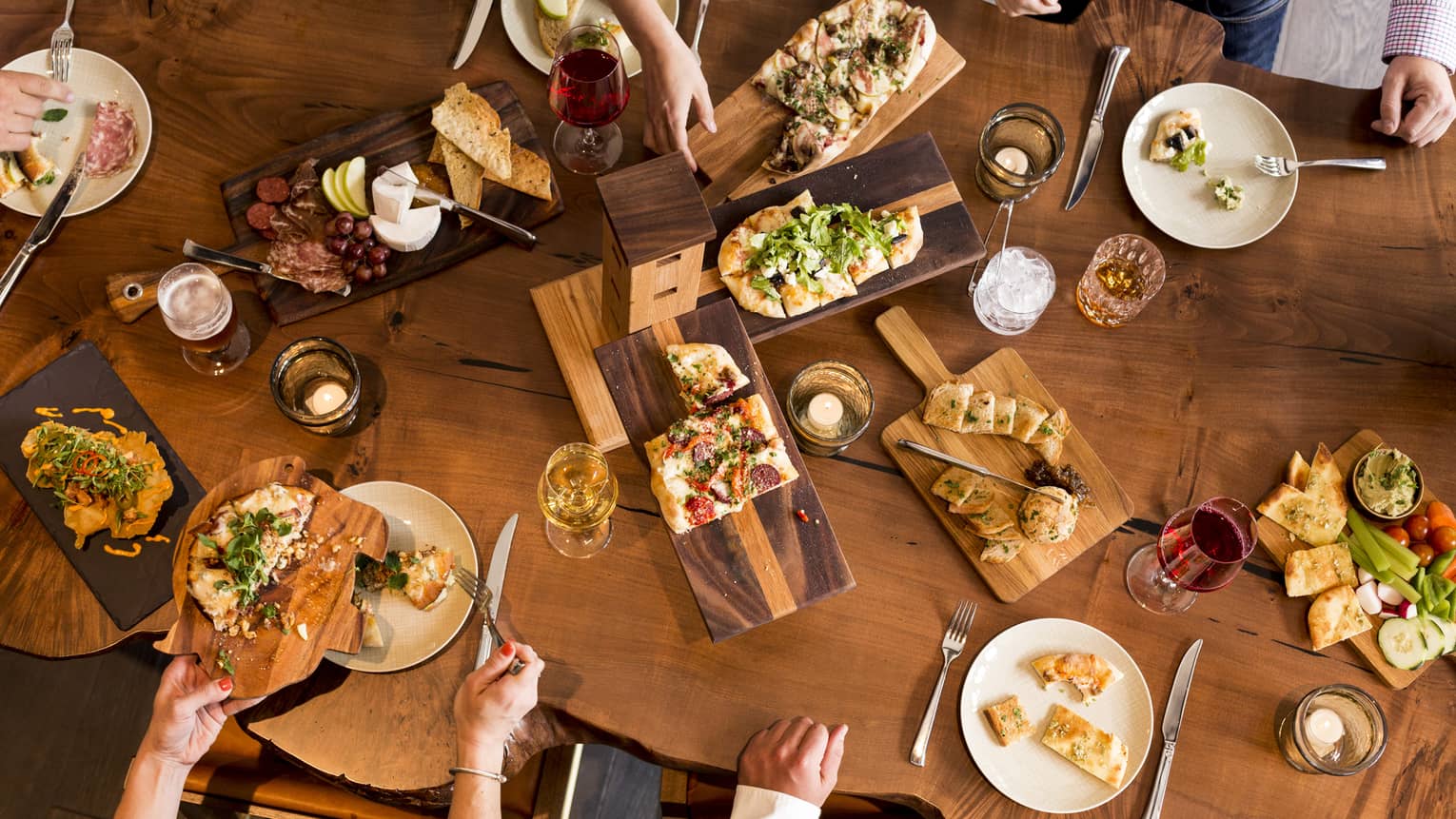 Aerial view of people dining around rustic wood table with platters of flatbreads, appetizers