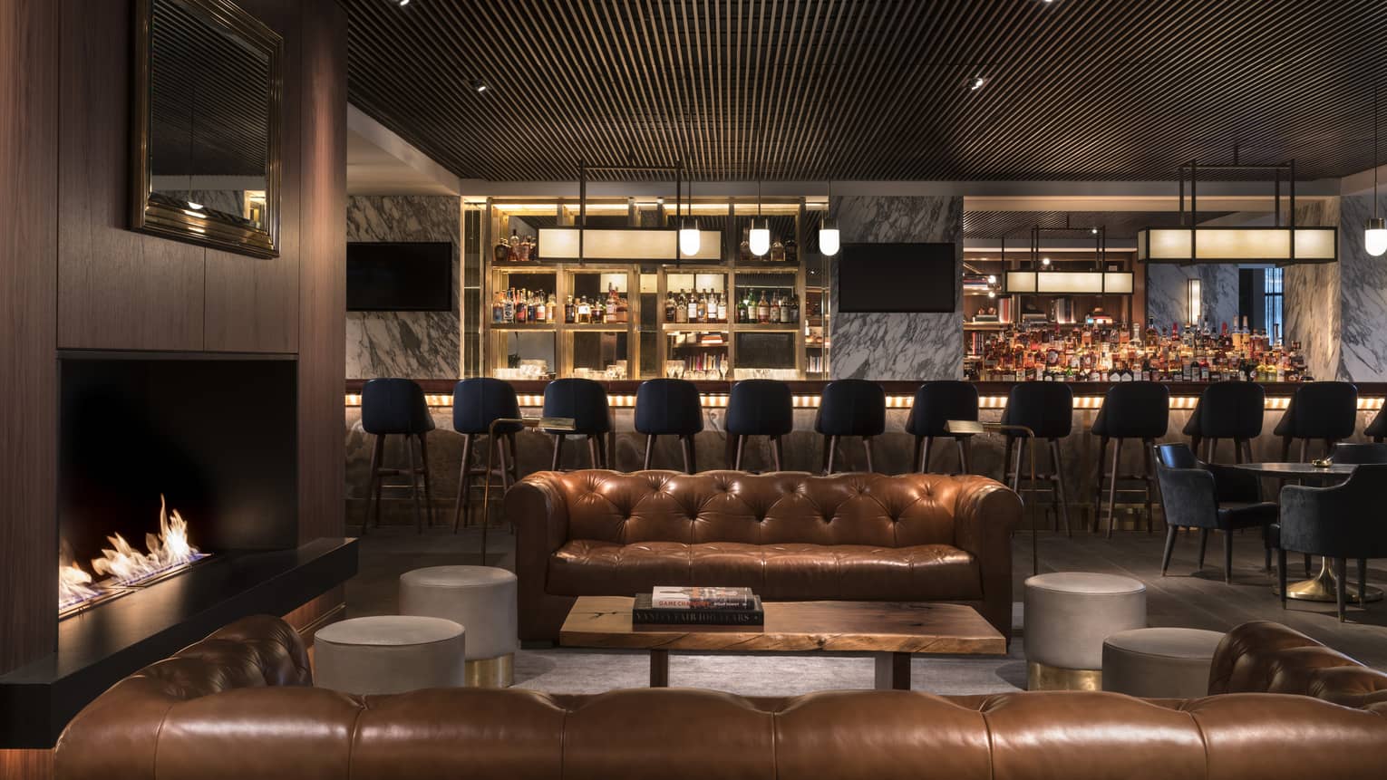 Two caramel-coloured Chesterfield sofas around a lit fireplace with cocktail bar in the background
