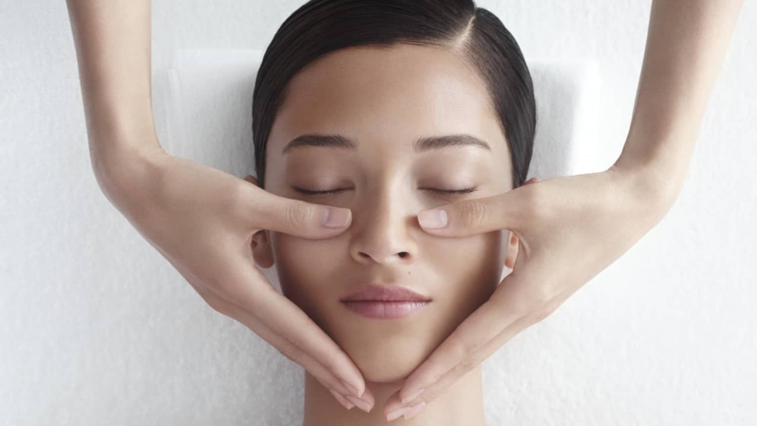Woman in white bathrobe lies on massage table with eyes closed, masseuse rests hands on her cheeks
