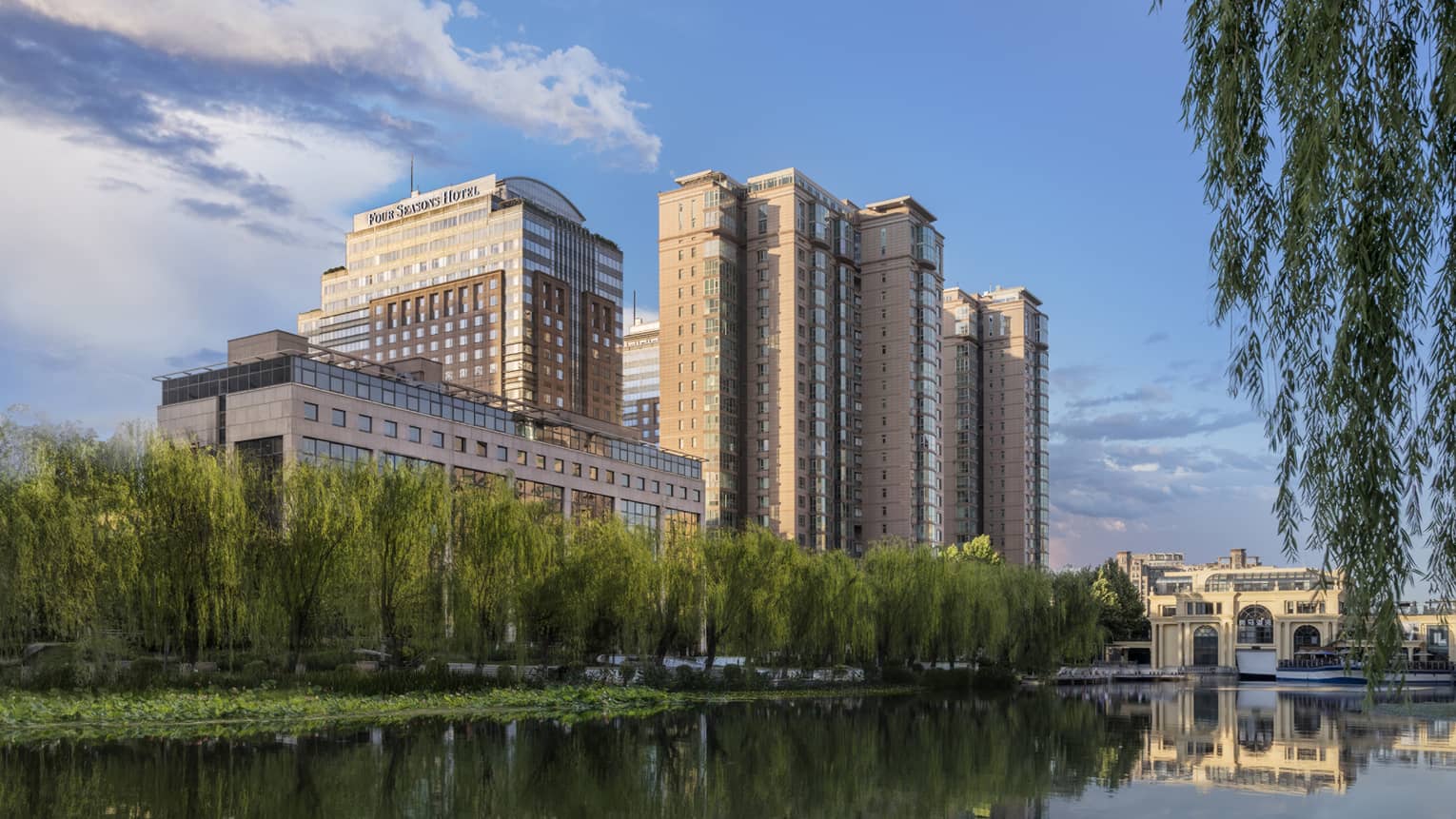 A large hotel building on the edge of a body of water, a line of tall green plants grows near the water.