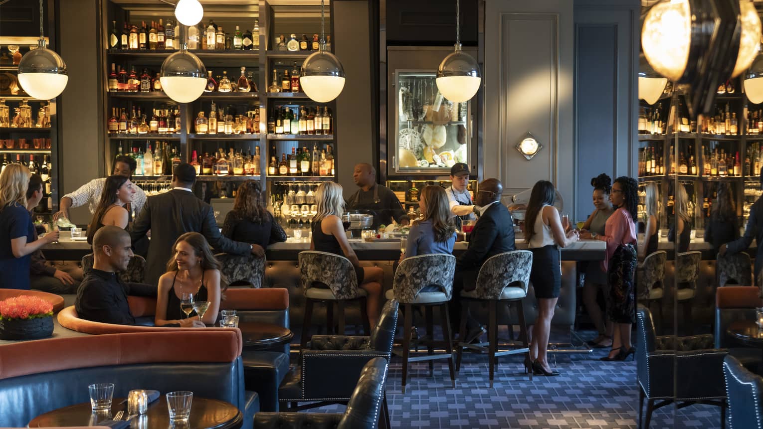 Guests and staff in a spacious, softly lit lounge space with both banquette and bar seating, hanging globe lights, and walls of shelves with bottles.
