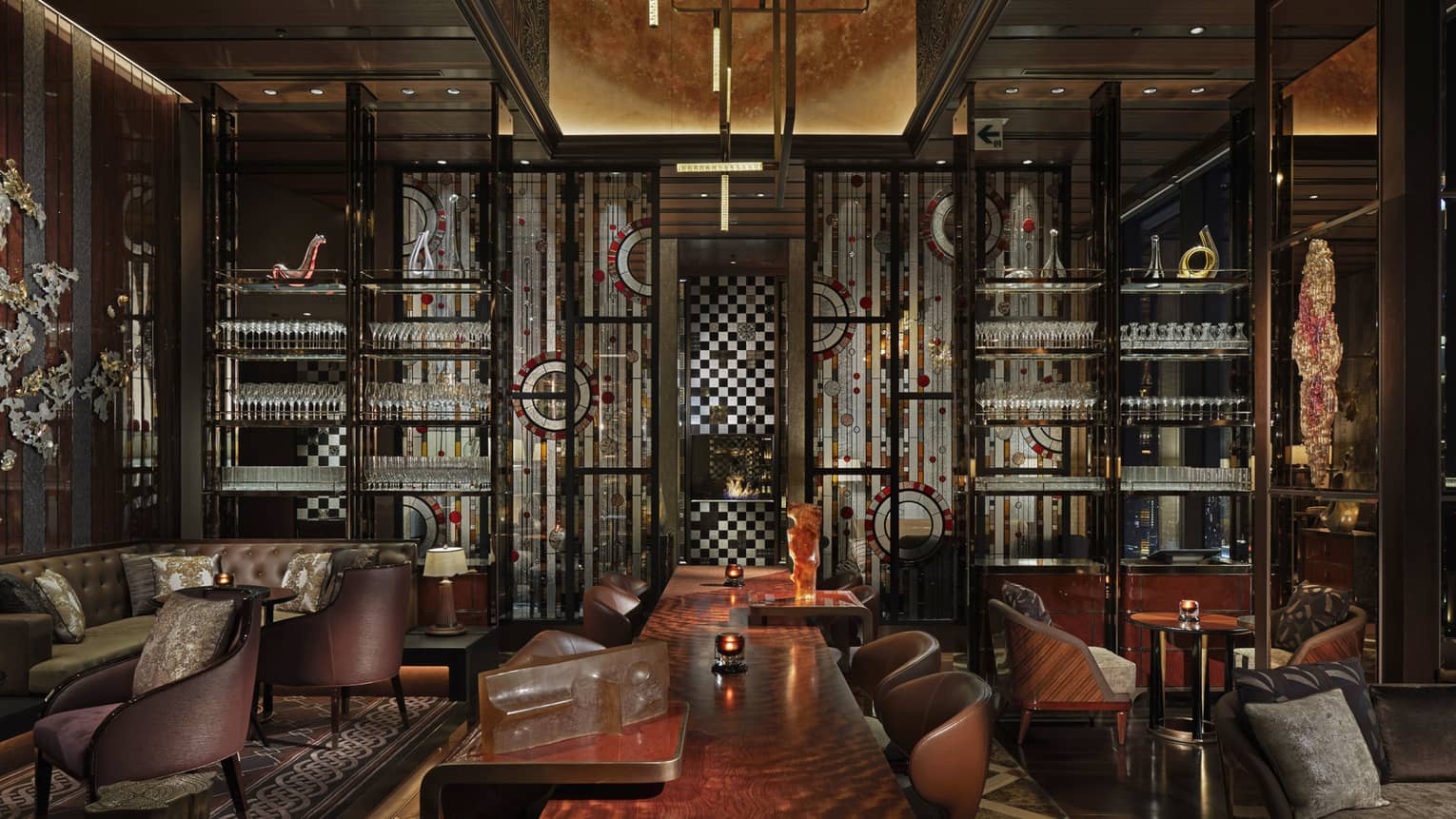 Lounge area with lounge chairs and ergonomic table in the center, shelves of cocktail glasses, lowlighting, muted tones