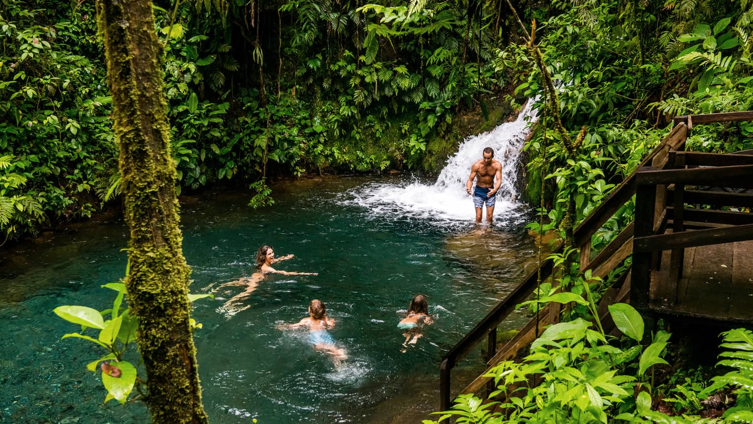 Two adults and two kids frolic at the foot of a waterfall surrounded by lush jungle greenery
