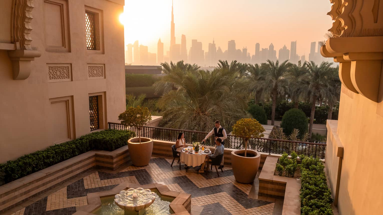 Table set for two on an open-air rooftop terrace with a chevron tiled floor and the sun rising over a city skyline in the near distance