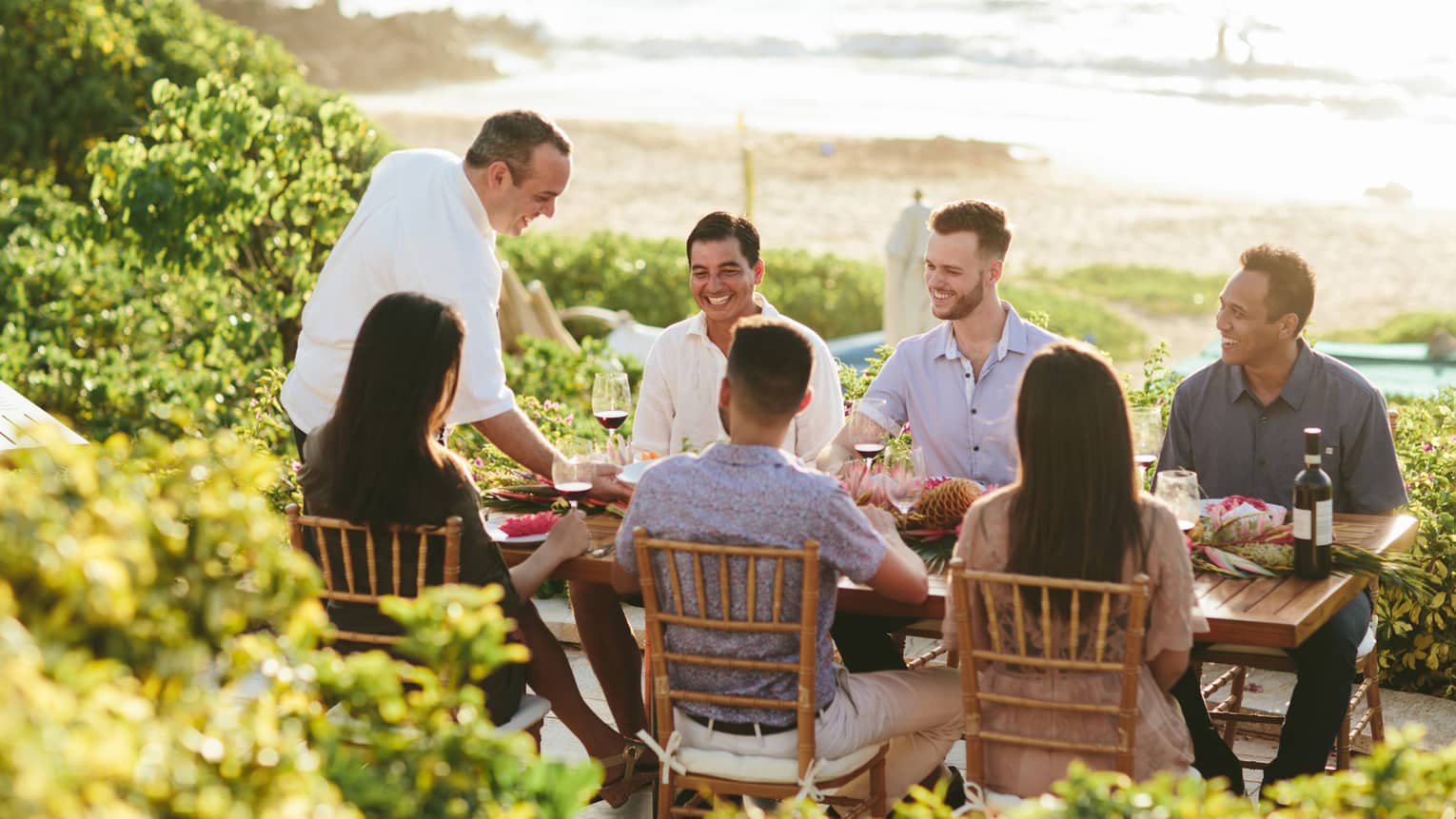 Six people smiling, sit around outdoor wooden table by ocean as wait staff serves them