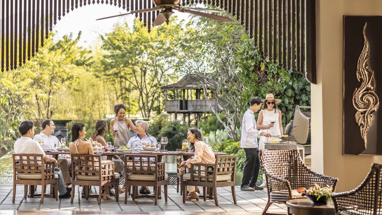 A long brown table with a family sat at it, in front of large trees.