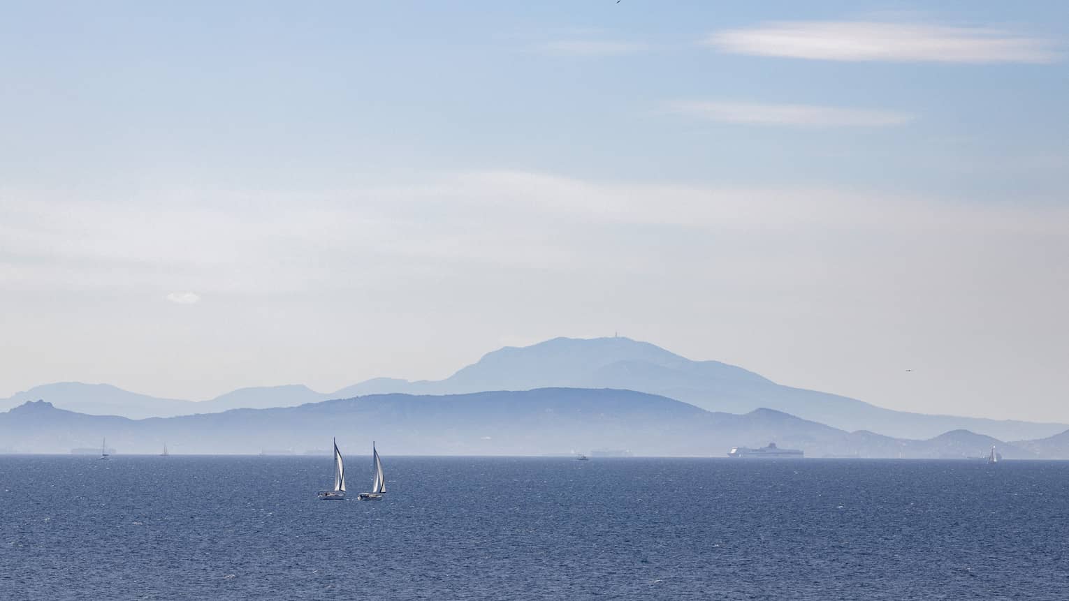 Two sailboats on blue ocean with blue-grey mountain range in the background 