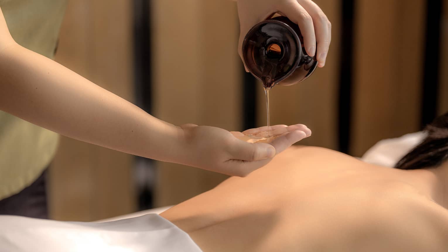Spa therapist pours oil into hand above woman?s bare back in preparation for a massage