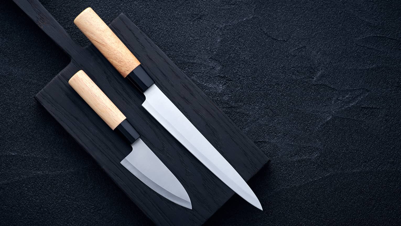 Two extremely sharp wood-handled knives, one long, one short, laid out on a black cutting board on a dark surface.