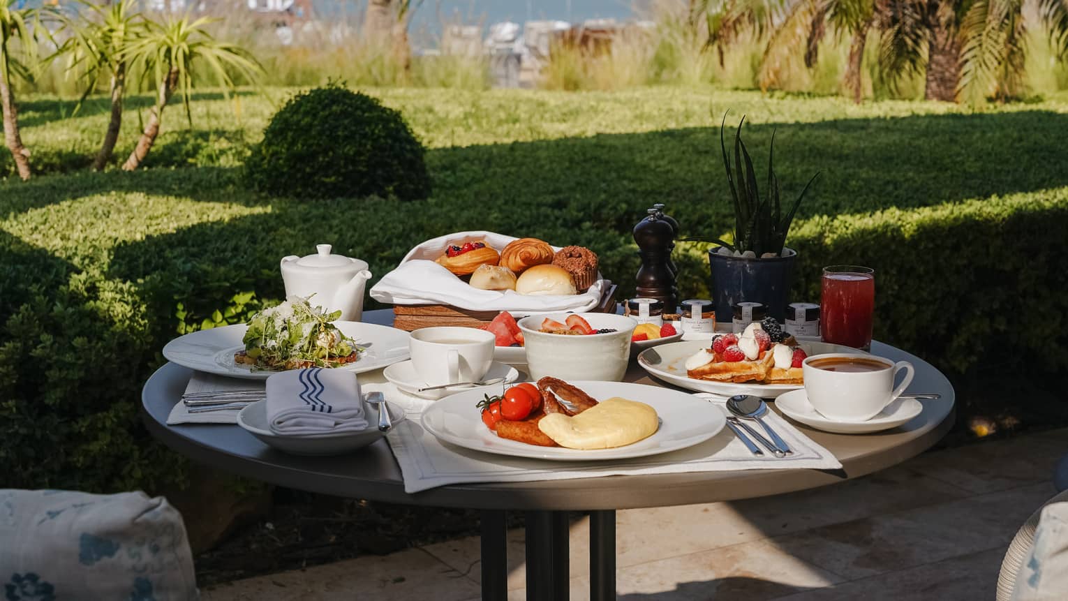 Outoor table filled with plates and bowls of breakfast food with a green lawn nearby and the ocean in the distance