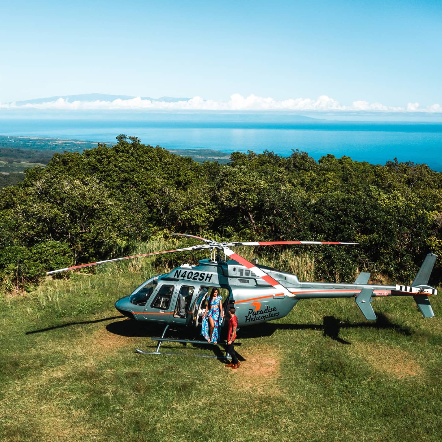 Woman and boy standing beside helicopter on grass with bushes and ocean water behind them