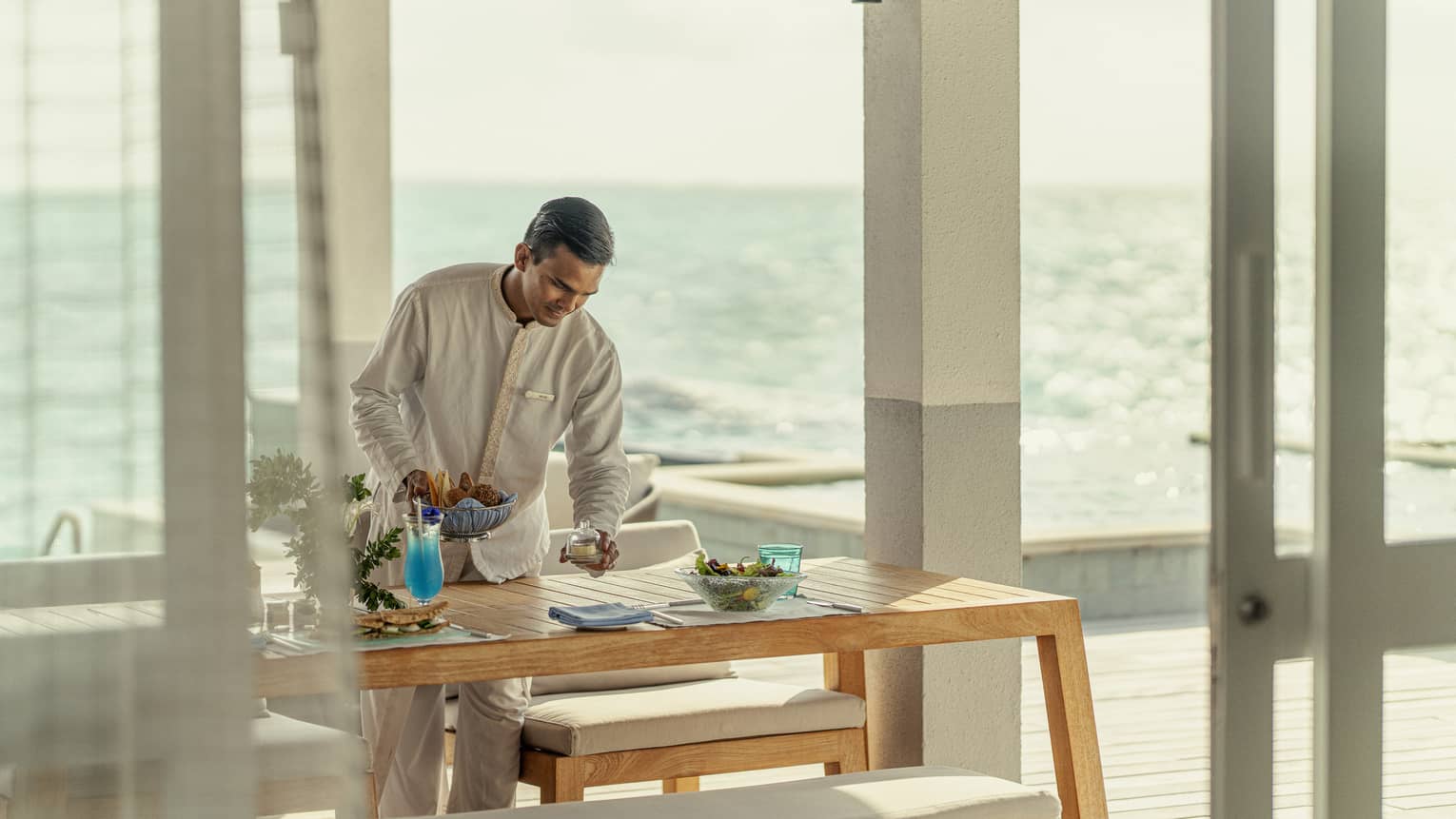 Male staff member sets table for In-Villa Dining, ocean in background