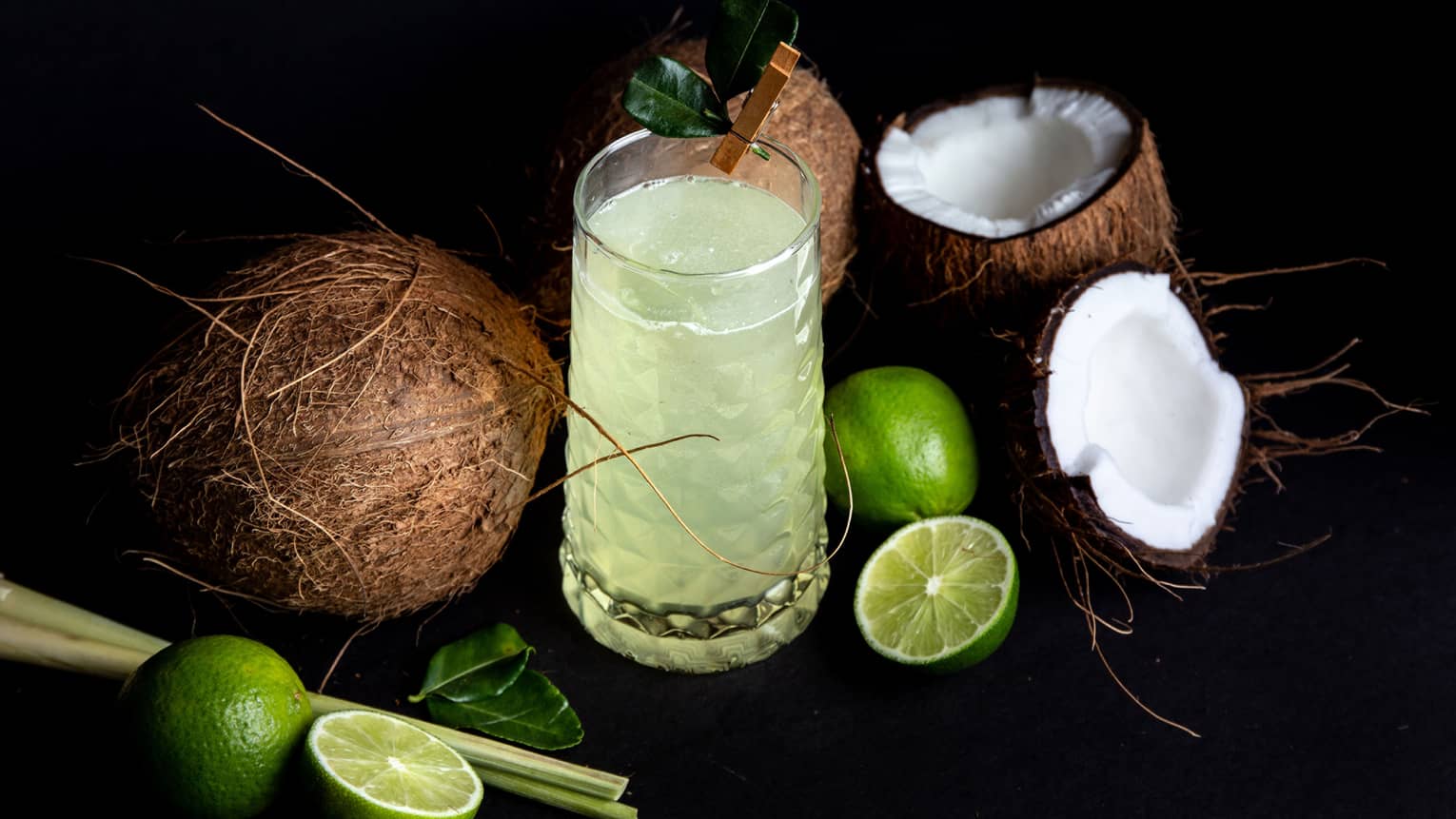 A tall glass of a light-green beverage sits in the middle of slived limes and two raw coconuts