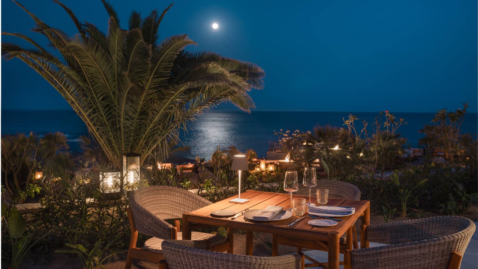 Dining table set for four on an outdoor patio at night with palm trees and the ocean in the distance