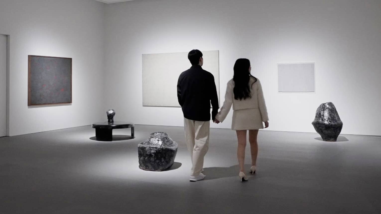 A couple holding hands walks through a modern art gallery with white walls, gray flooring and spotlights illuminating the art pieces