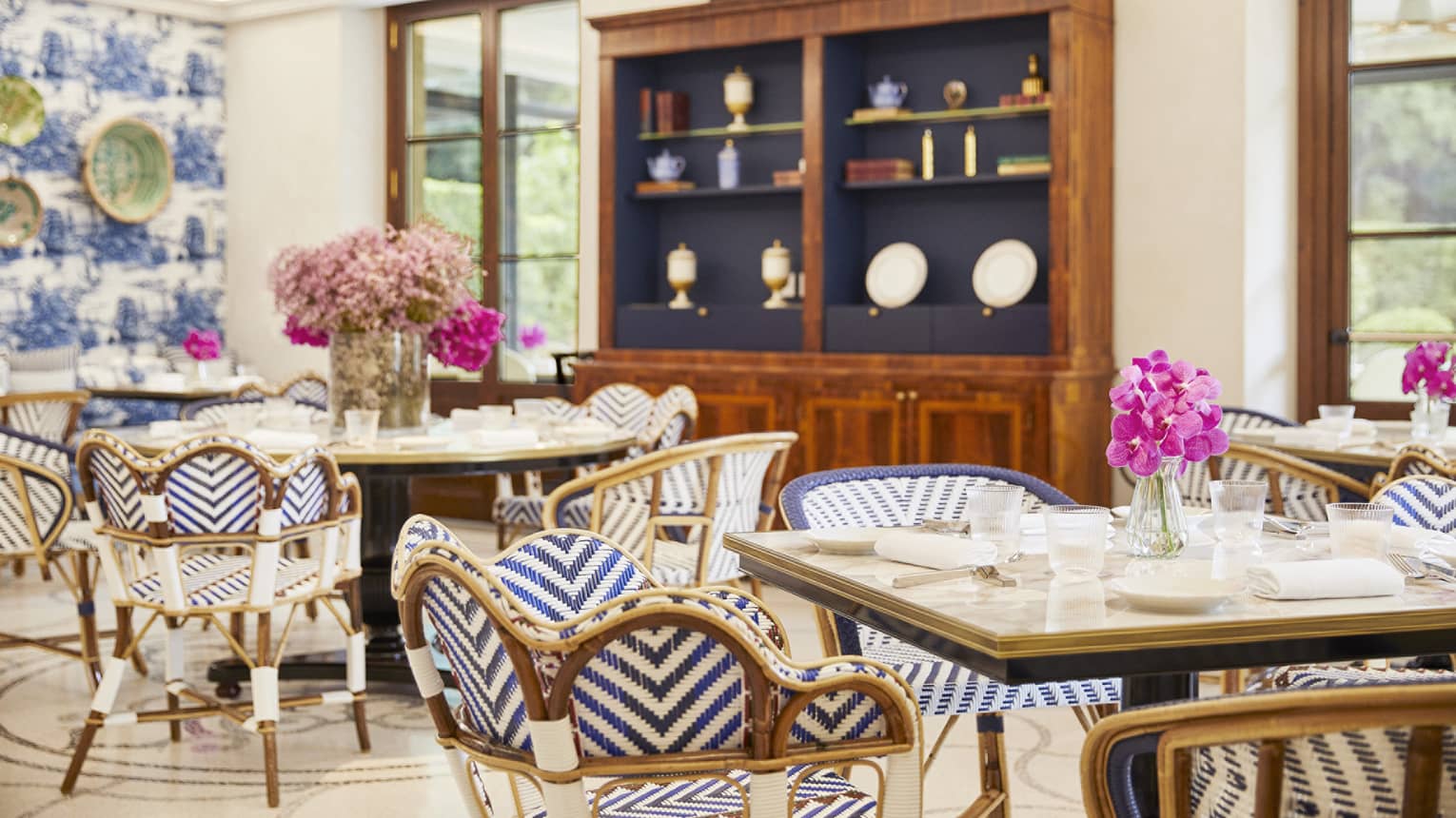 A bright, spacious indoor dining space with dining tables, blue and white striped chairs, large windows, overhead lighting, and a large wood display cabinet next to one wall