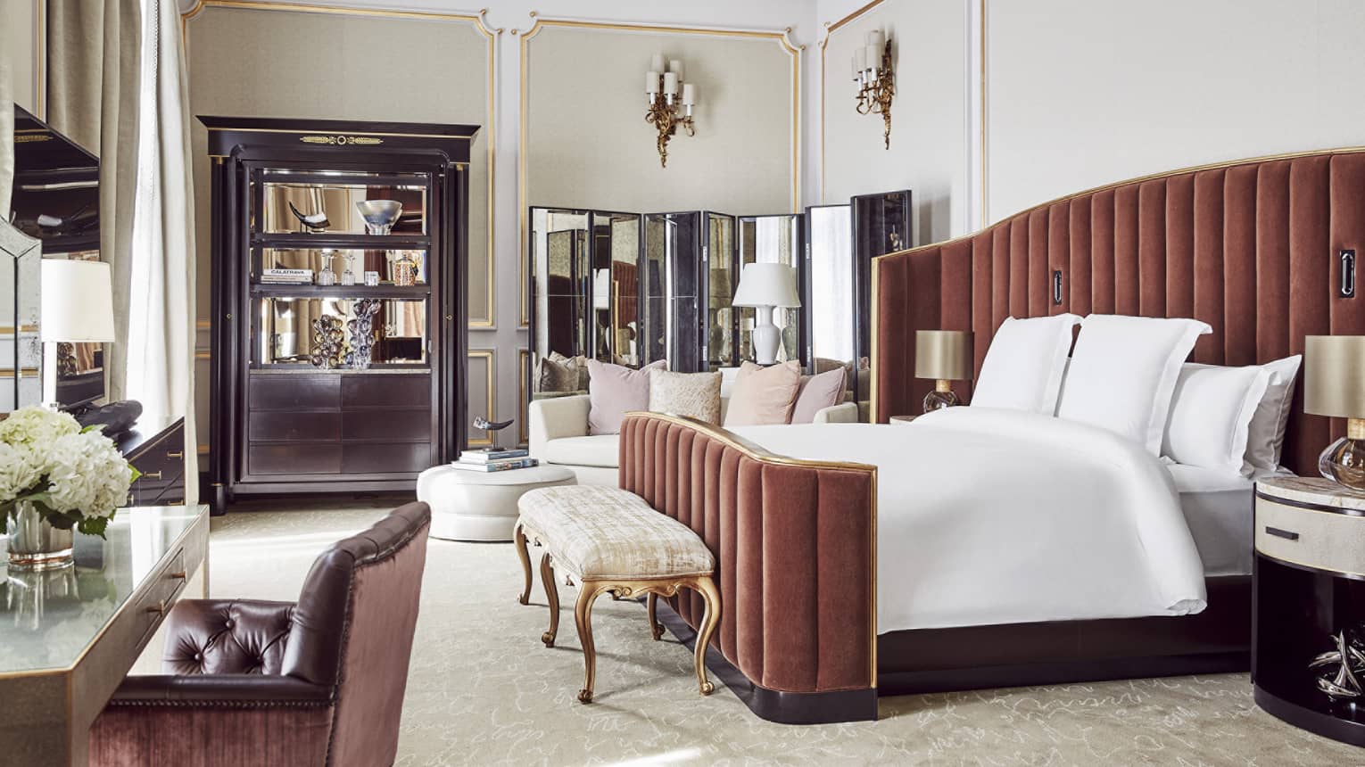 Hotel bedroom with a king bed with tall, upholstered headboard, gold-accented bench, sofa, ottoman, desk, mirrored divider, decorative cabinet and wall sconces