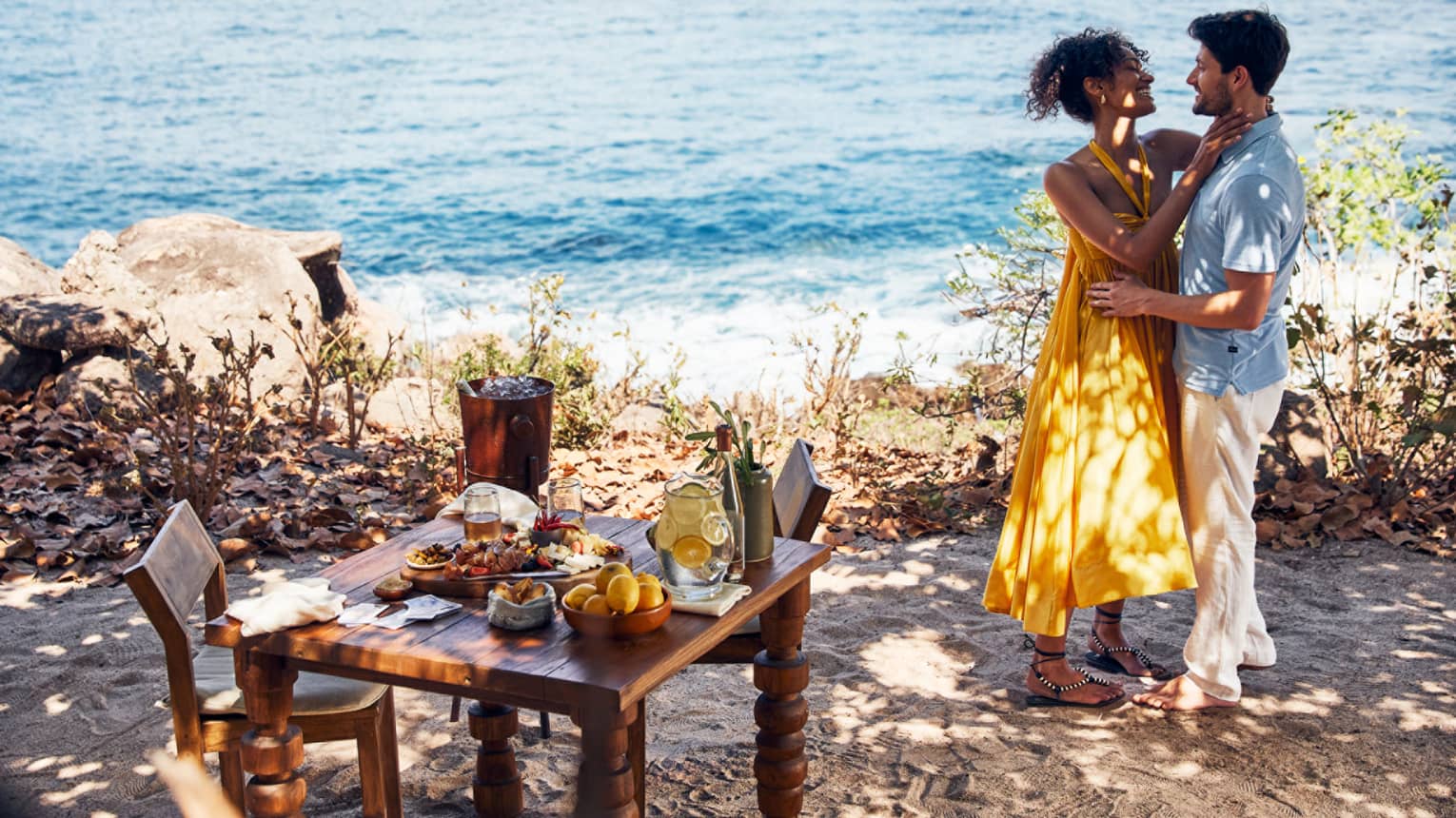 A man and woman dancing near the ocean with a small table of food near them.