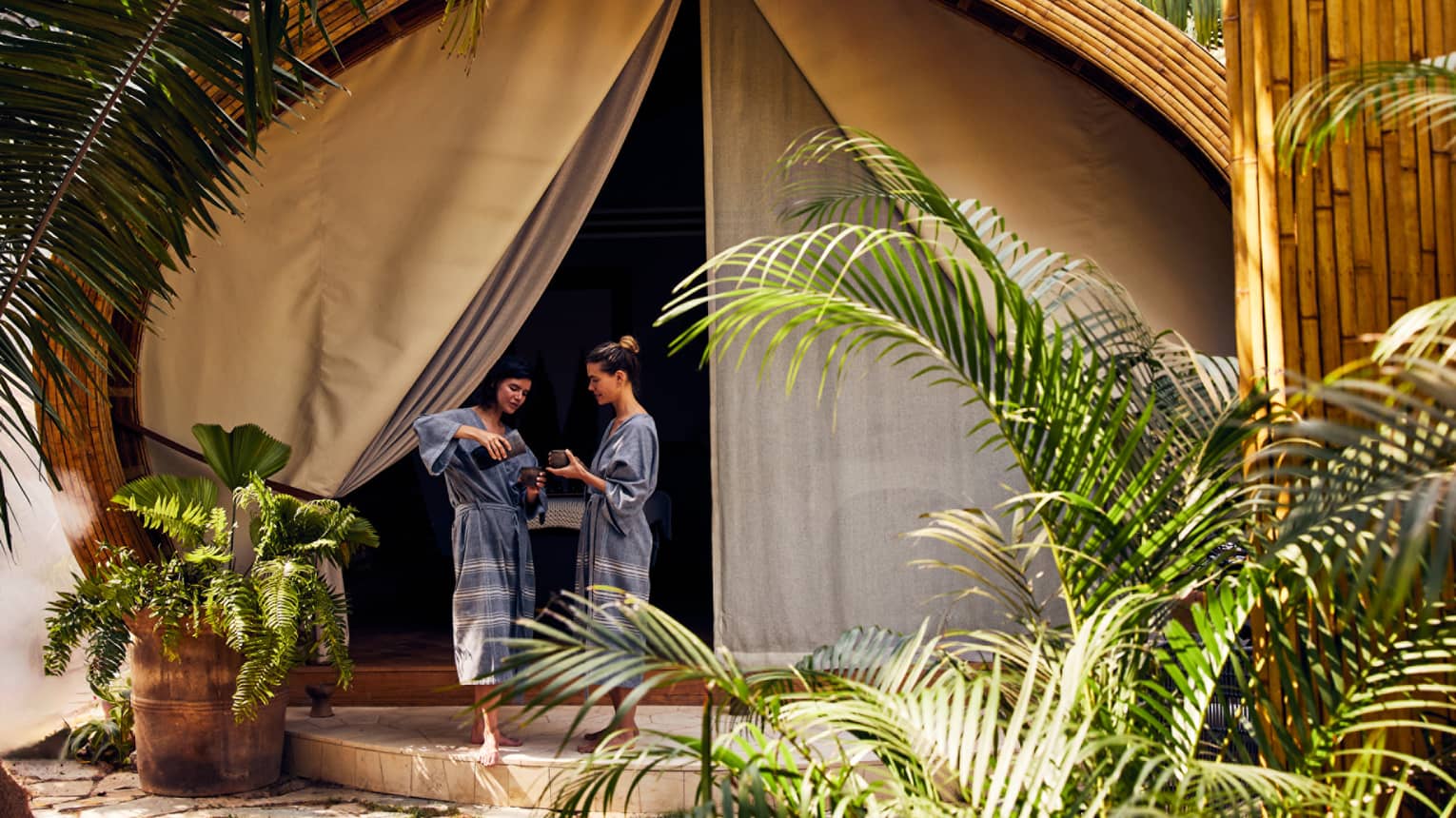 Two women in spa robes stand outside a luxury spa tent, one pouring tea for the other, surrounded by lush greenery in a peaceful setting.