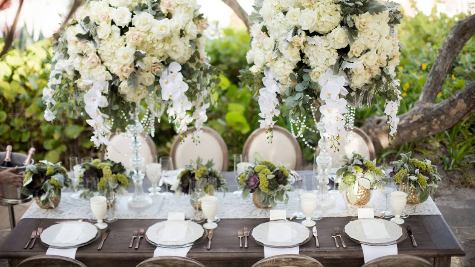 Outdoor banquet dining table under large white rose flower arrangements with hanging orchids