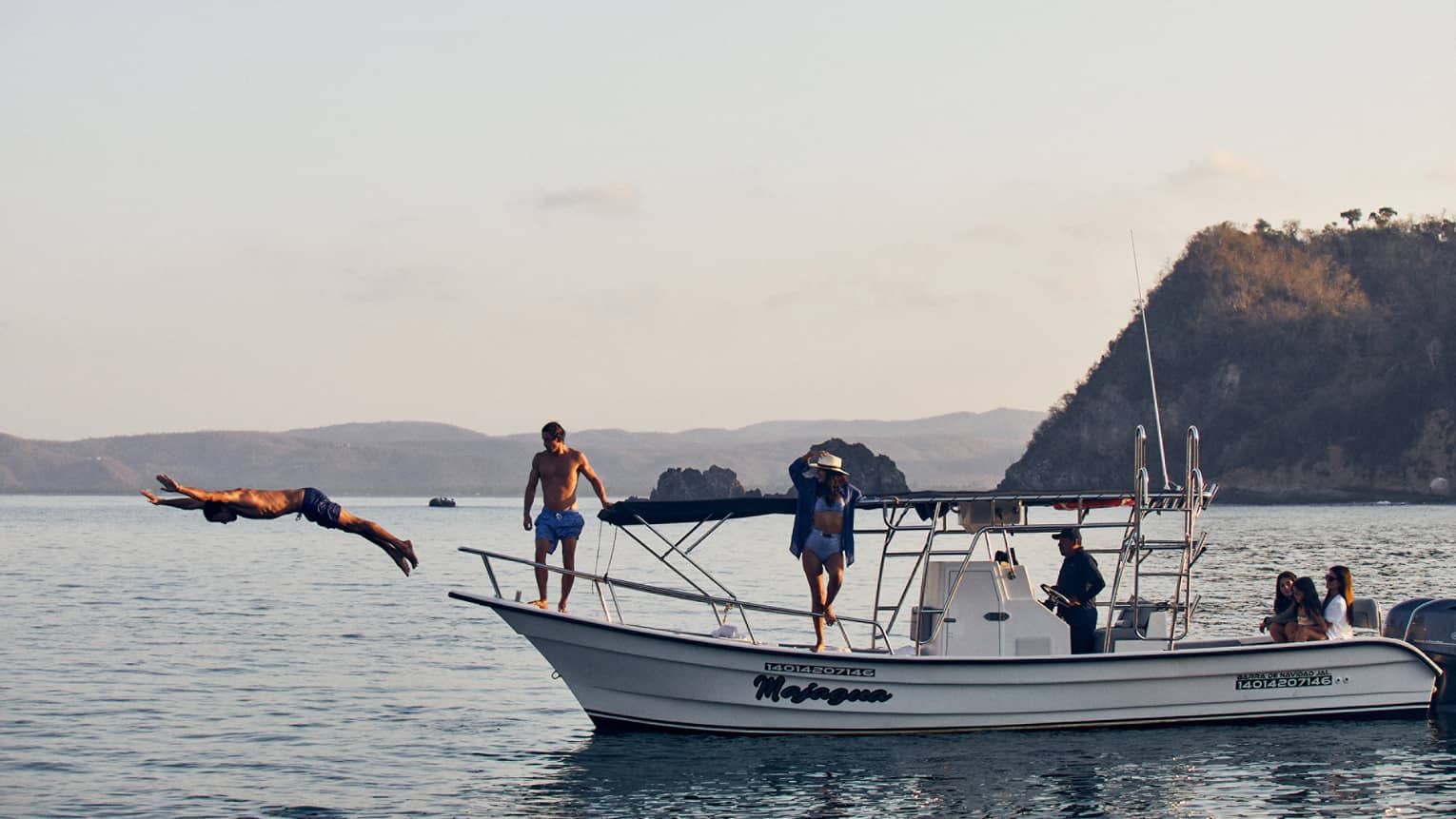 Side view of a guest diving gracefully into the water while others watch from a boat's deck, distant landmasses beyond.