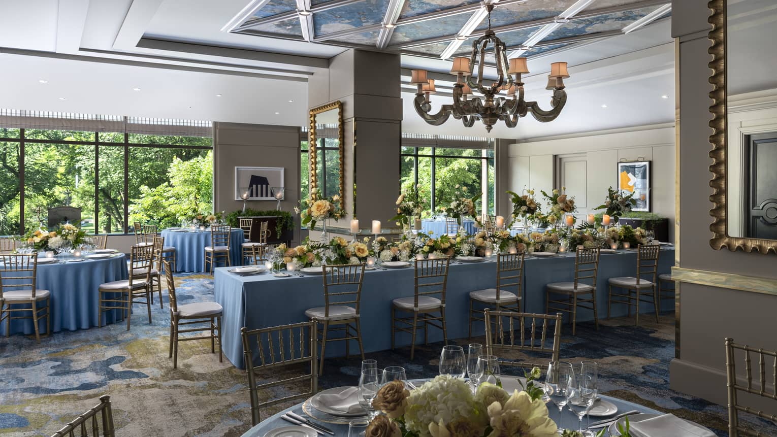 Long and round tables with blue tablecloths with yellow flowers, an old looking chandelier hangs from the ceiling.