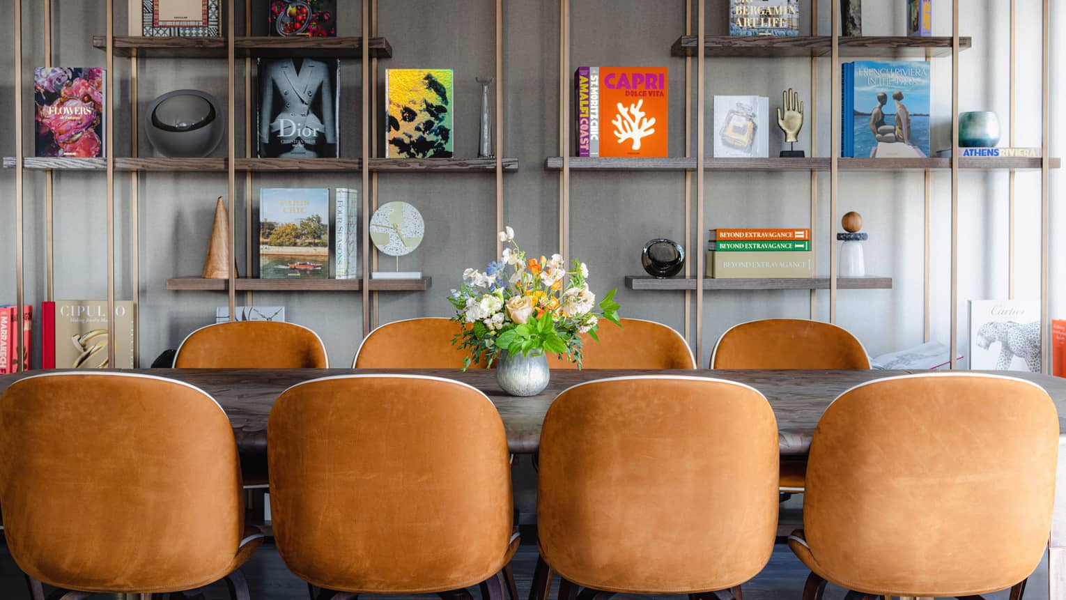 Rectangular table with floral centerpiece and eight suede chairs, copper bookshelf with books and trinkets