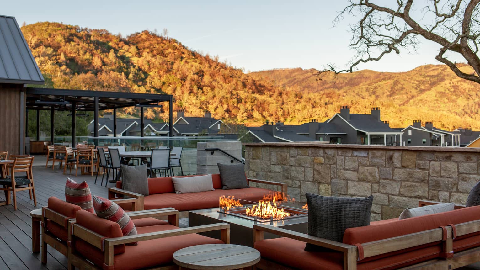 Pillowed couches around a fire table on an empty patio beside quaint cabins and hills with orange trees under a clear sky.