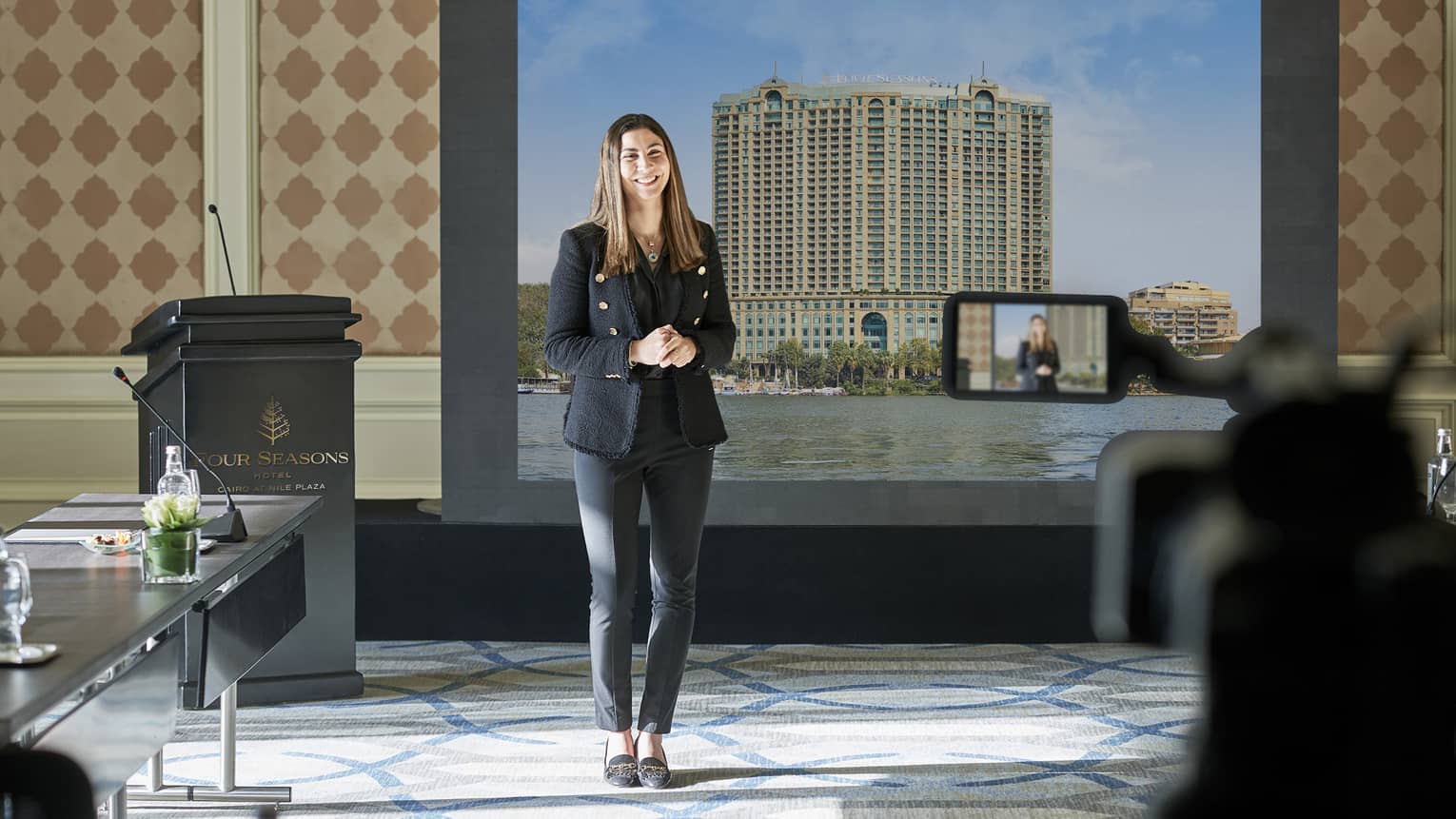 Standing woman presenting in a hybrid boardroom setup, with large screen in background.
