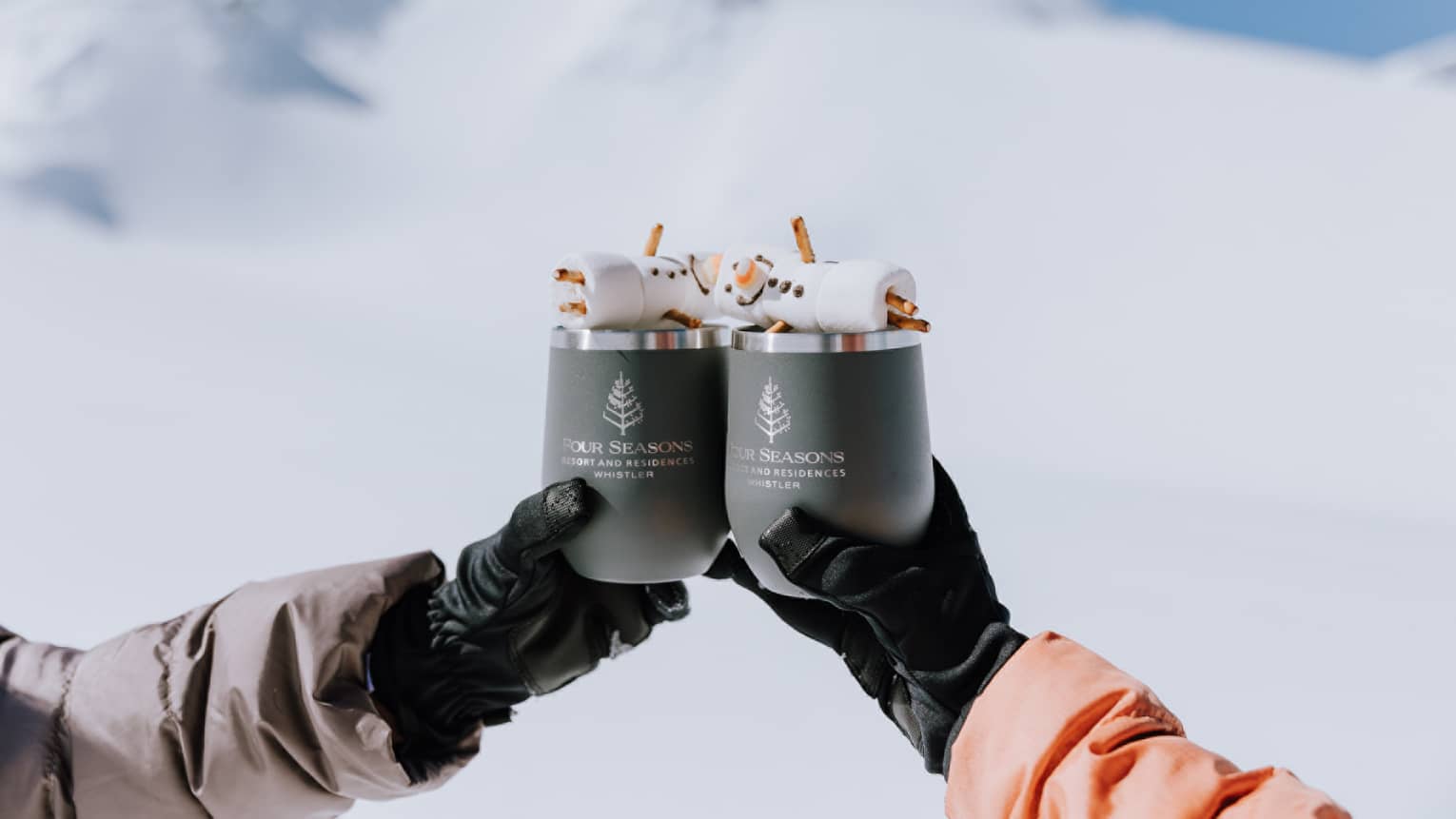 Two gloved hands raise insulated mugs of hot chocolate in a toast, snowy mountains in the background