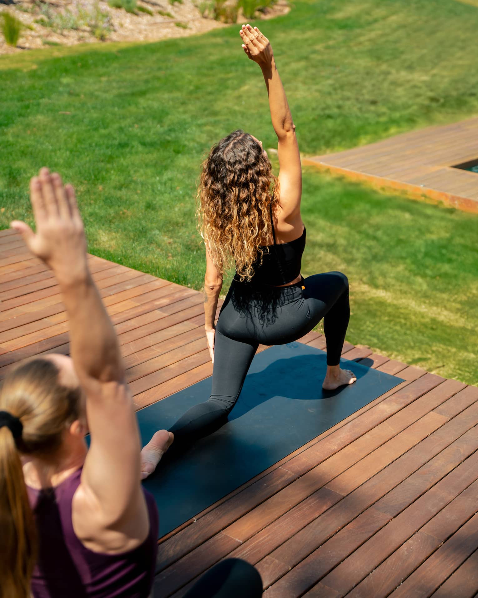 Two people in yoga pose practise on outdoor wooden deck overlooking lush green grass