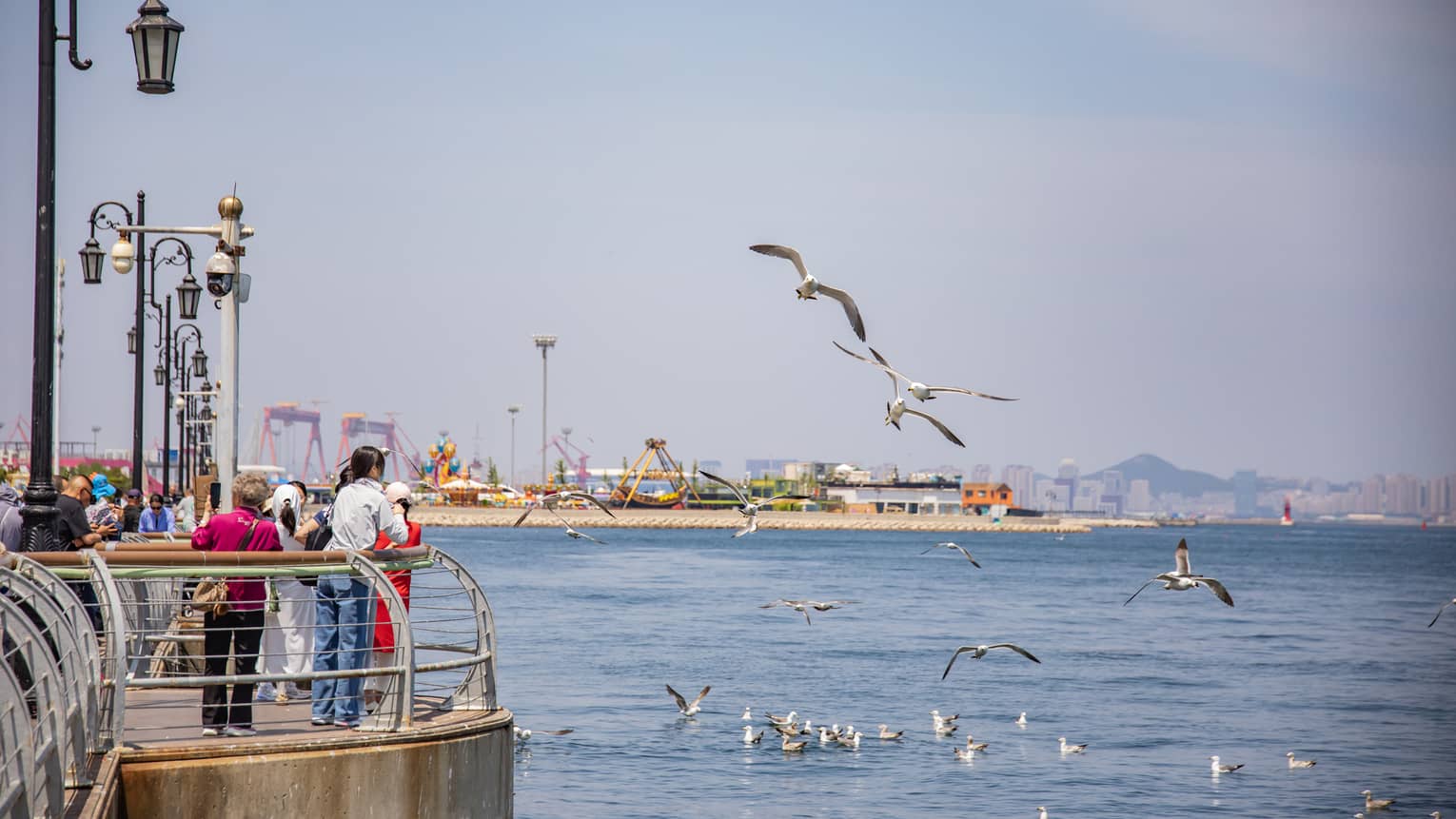 Tourists gather at a sunny pier lined with old-style lamp-posts as seagulls fly nearby, an amusement park in the distance.