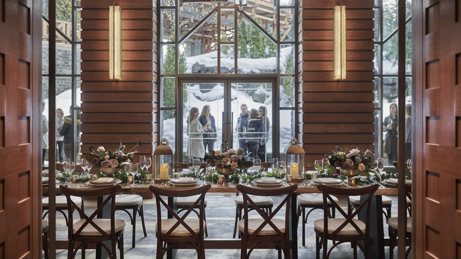 Elegant winter wedding setup inside Four Seasons Whistler, viewed through glass doors leading to a cozy, beautifully decorated dining area. The room is filled with wooden tables set with fine dinnerware, crystal glassware, and lush greenery centerpieces, all illuminated by warm overhead lights. Outside the glass doors, guests in formal attire chat in a snowy courtyard, adding to the festive and romantic atmosphere.,Elegant winter wedding setup inside Four Seasons Whistler, viewed through glass doors leading to a cozy, beautifully decorated dining area. The room is filled with wooden tables set with fine dinnerware, crystal glassware, and lush greenery centerpieces, all illuminated by warm overhead lights. Outside the glass doors, guests in formal attire chat in a snowy courtyard, adding to the festive and romantic atmosphere.