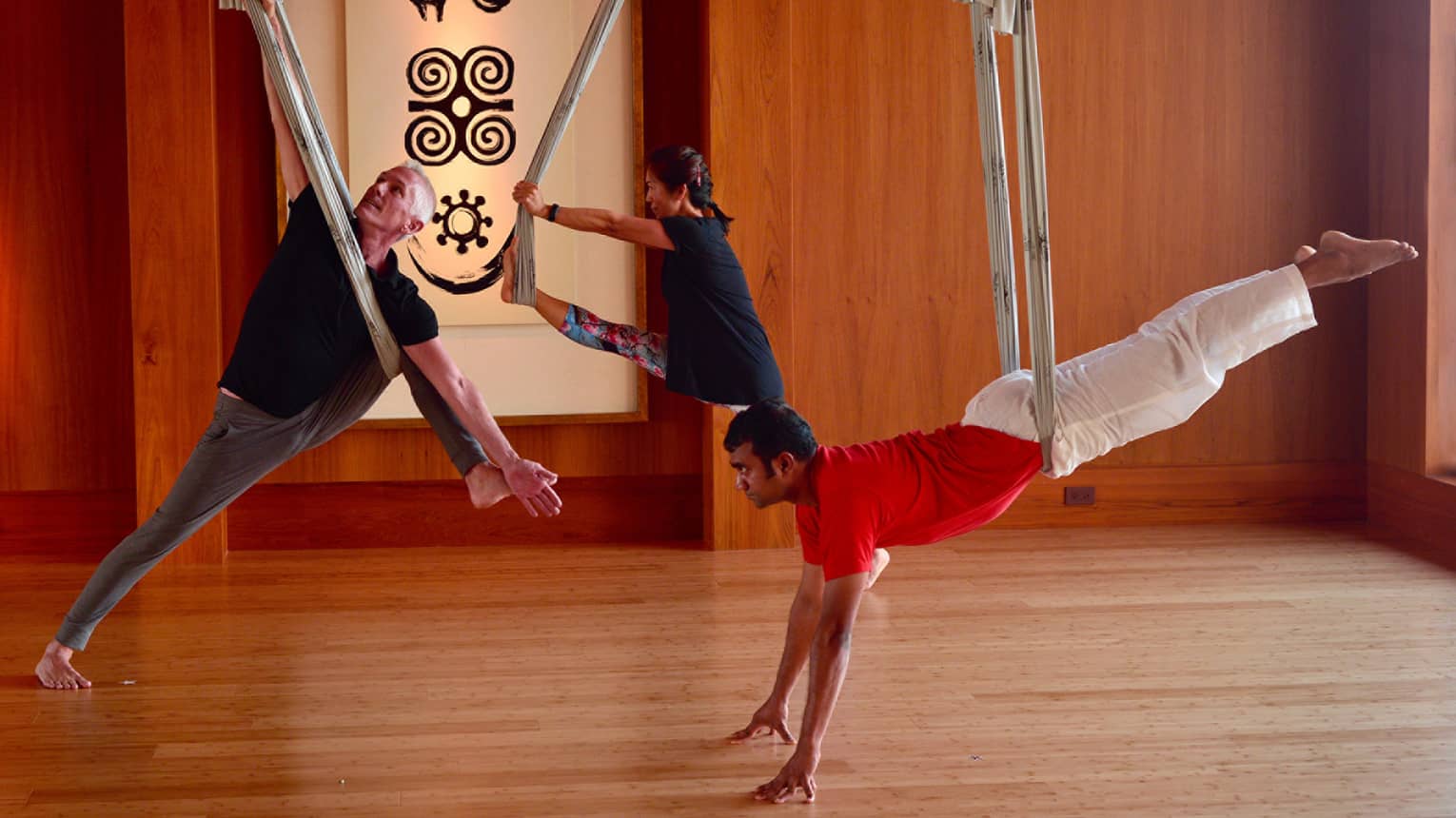 Three people balance on fabric aerial yoga hammocks hanging from ceiling