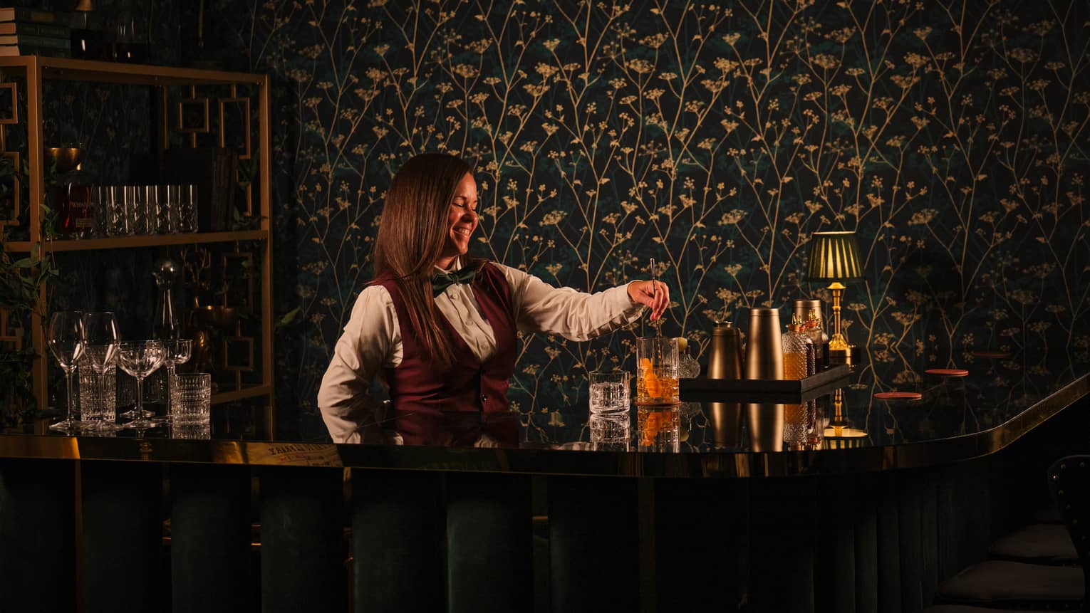 Bartender wearing white shirt, burgundy vest and black bowtie pours a cocktail from behind the bar