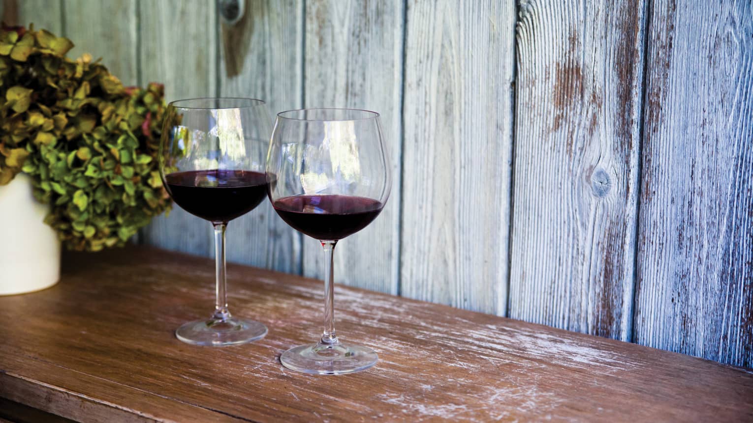 Two glasses of red wine and sit on wooden table, with white vase filled with hydrangeas