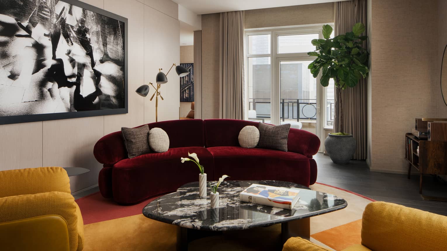 Luxury suite living room with burgundy sofa, coffee table, two yellow armchairs, large black-and-white wall art, potted plant and city-view balcony
