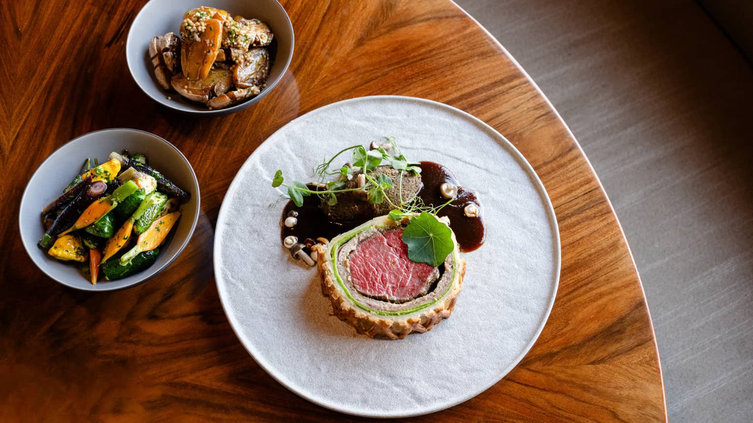 Plate of beef Wellington set next to a bowl of vegetables and a bowl of mushrooms on a round wooden table
