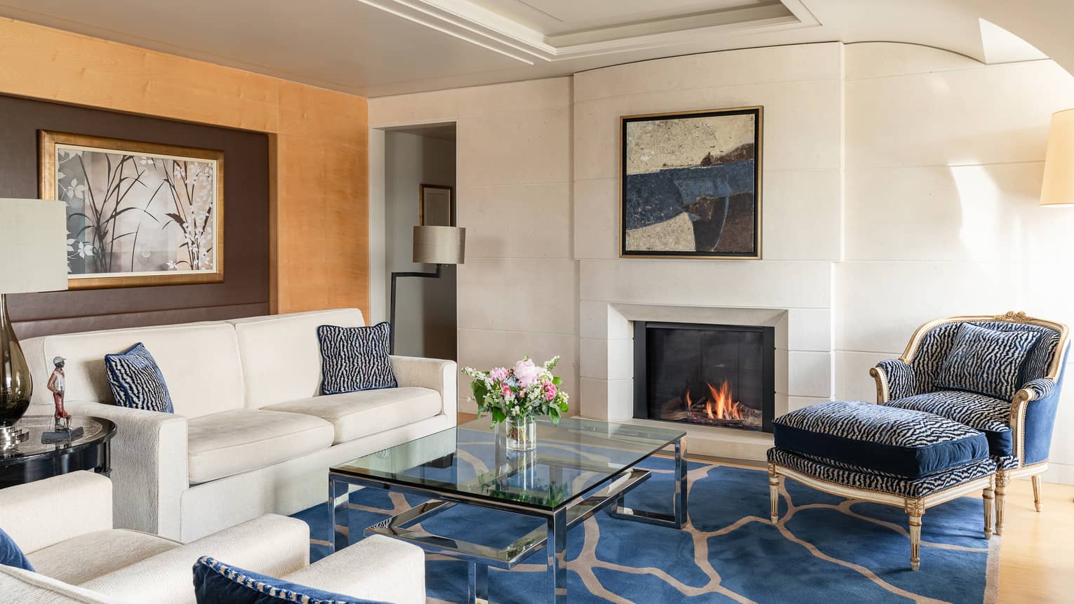 Luxury hotel suite living room with a cosy fireplace, cream sofa, blue and cream armchairs, glass coffee table, floral arrangement and wall art