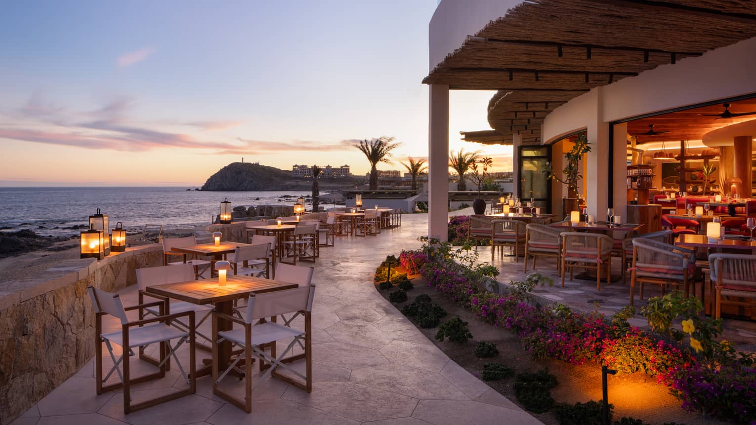 In the warm afterglow of sunset, lanterns light up a large, curved stone restaurant patio at the edge of the ocean.