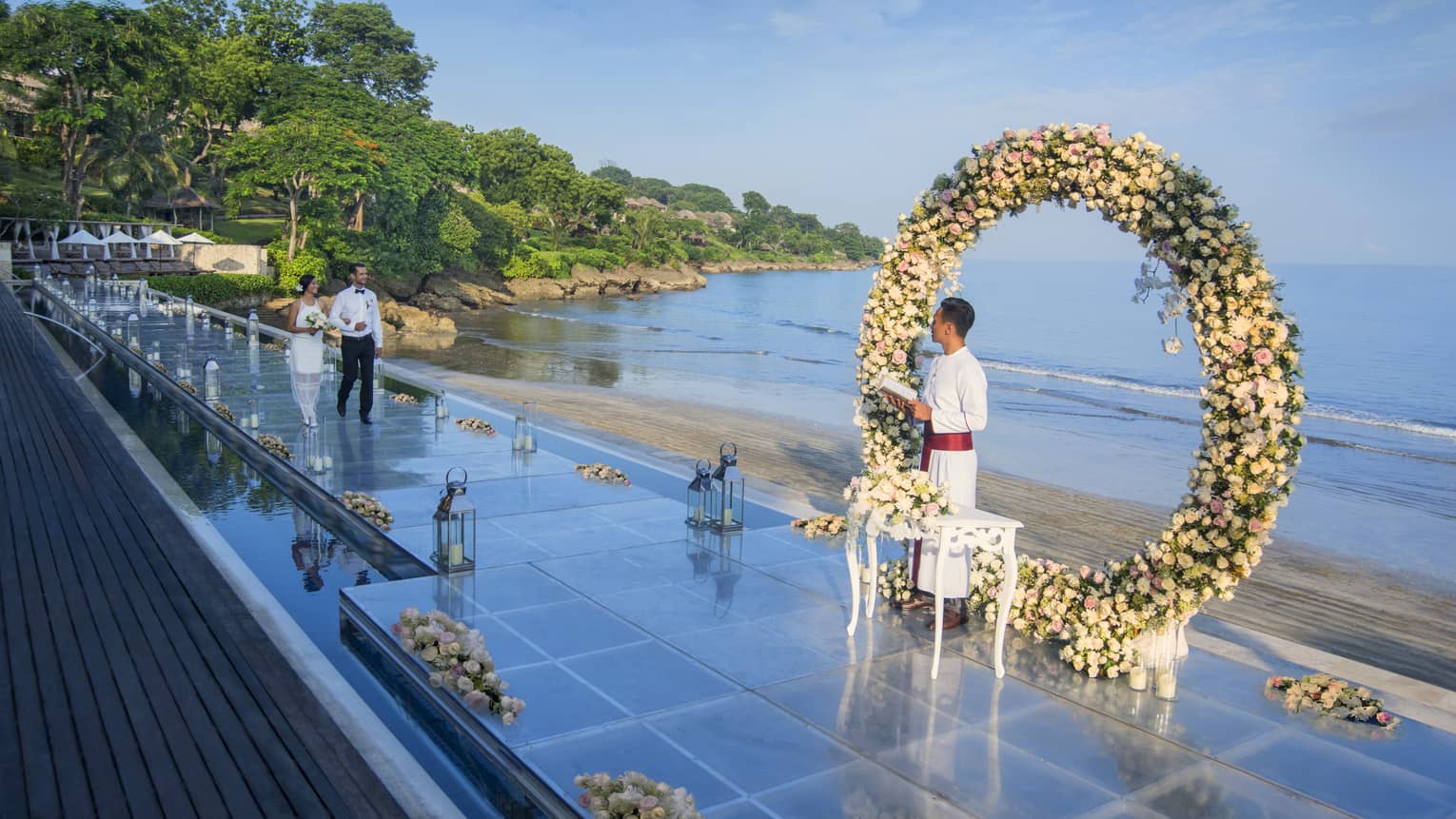 A couple walks down the aisle toward a flower arch during an outdoor wedding ceremony