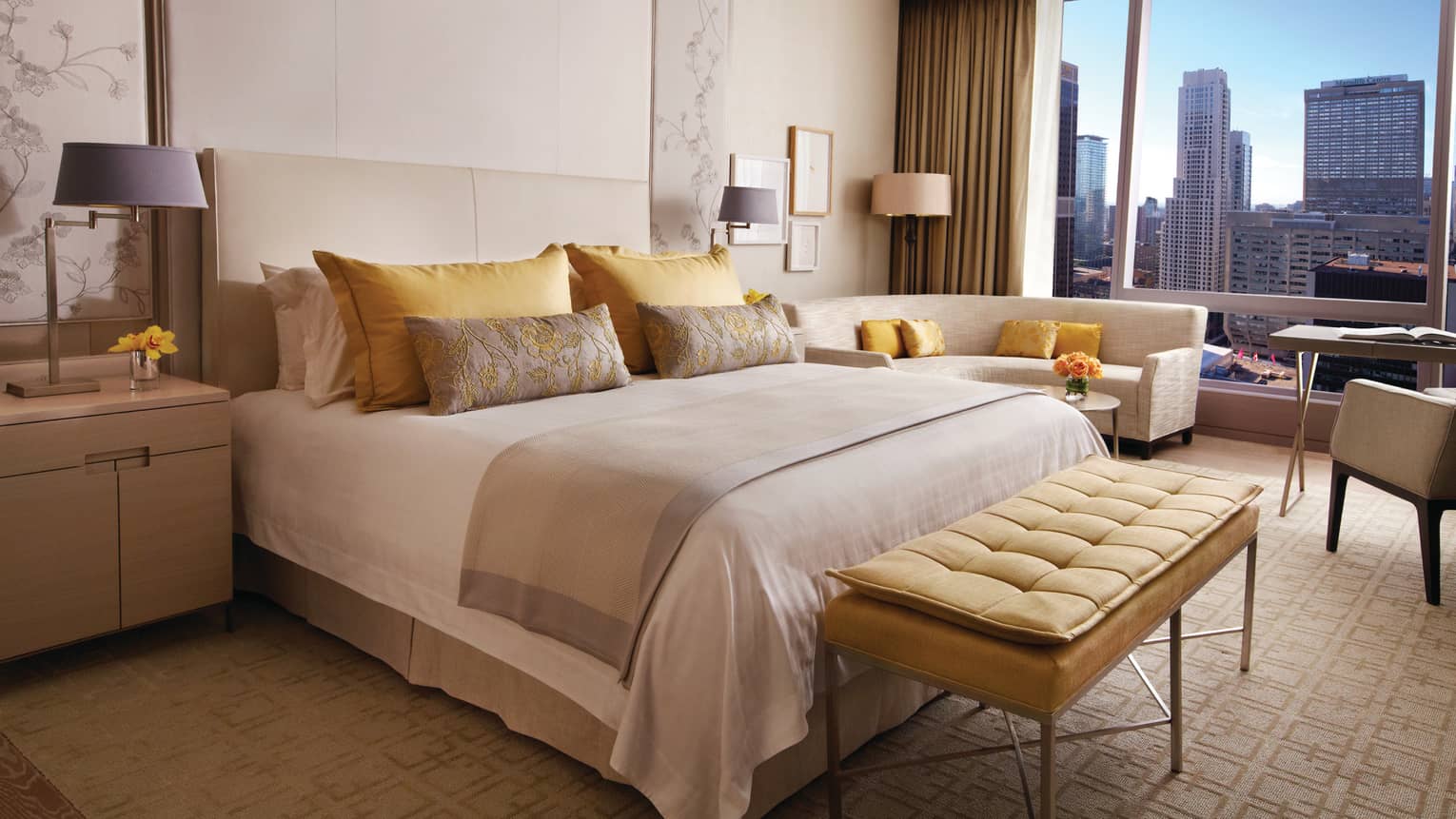 Plush yellow bench at foot of bed with yellow, gold pillows by curved loveseat, window