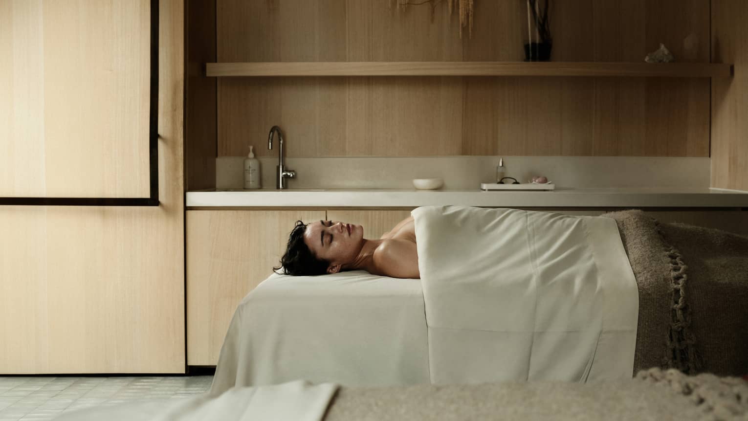 A person lies on supine on a spa treatment table in a serene, spacious treatment room and is draped with with a sheet and blanket.
