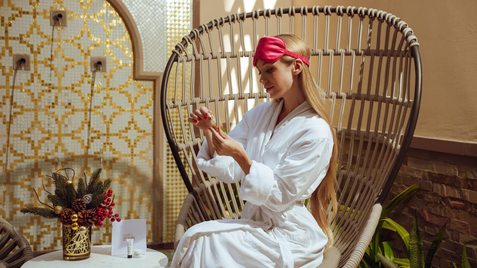 Person with waist-length blonde hair wears a white spa robe and a hot pink satin eye mask on their head while sitting on a rattan butterfly-style chair and painting their nails