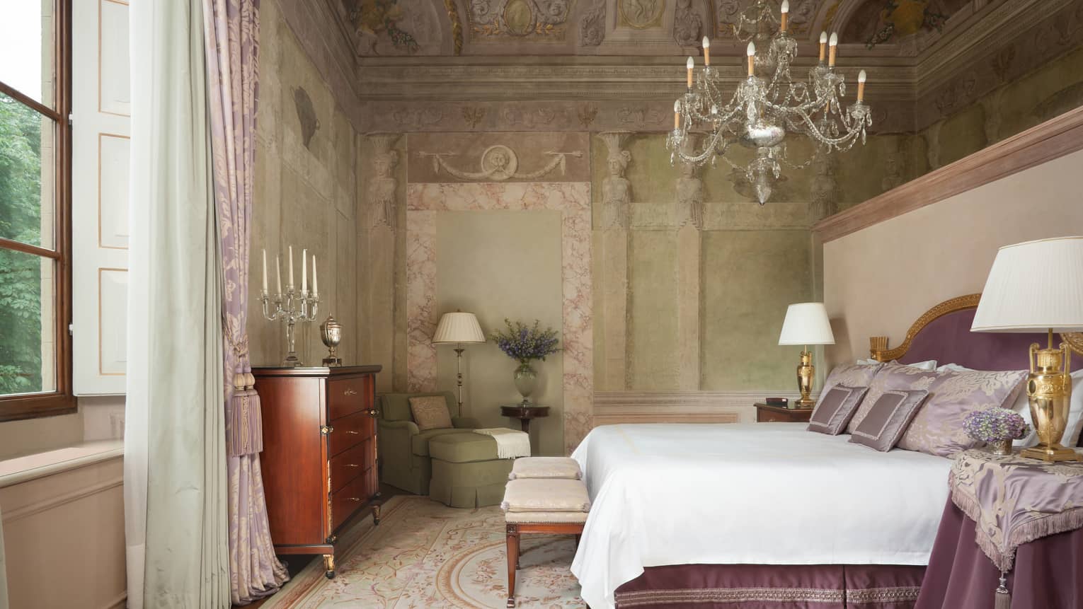 Frescoed Executive Suite bed with purple velvet headboard, nightstand with cloth under chandelier, vaulted ceiling